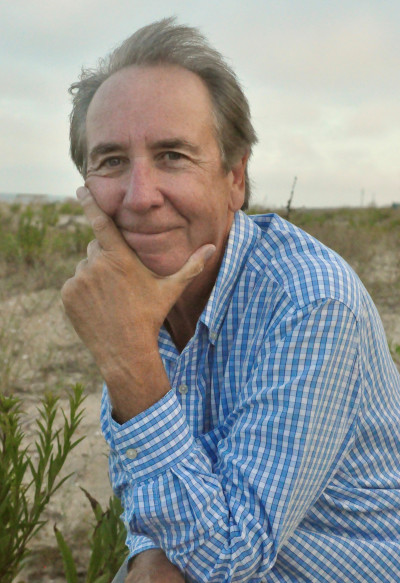 Headshot: Thomas Guay, a man with windblown hair wearing a blue checkered shirt with his chin in his hand, smiling at the camera, with sand and grasses in the background.
