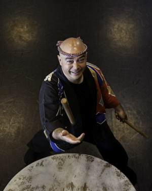 Mark Rooney stands behind a Taiko drum, smiling and tossing a drumstick into the air. He is wearing traditional Japanese dress for drumming.