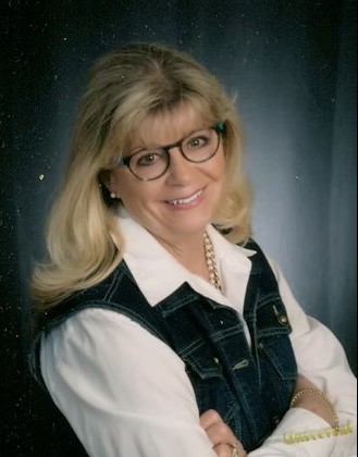 Headshot of a woman with long blond hair, wearing glasses, a white colored shirt and black vest, smiling at the camera