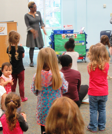 A group of children and adults, some sitting and some standing, watching a storytime presenter.