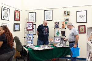 male artist behind a table displaying his artwork and another male speaking to him