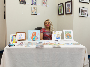 female artist sitting behind a table with artwork on the table and hanging on the wall