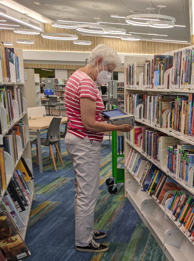 Woman wearing a mask holding a tablet, looking at a shelf of books