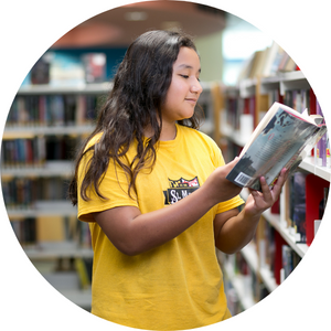 Teen girl with yellow shirt and brown hair standing at a book shelf reading a book