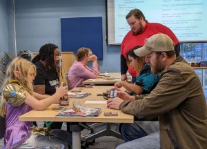 Three adults and a young girl play a tabletop game while another man stands next to the table