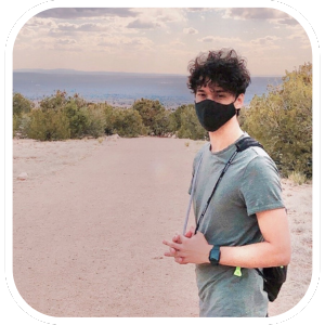 Photo of a young man wearing a green tshirt and a black face covering standing in foreground against a backdrop of trees and mountains