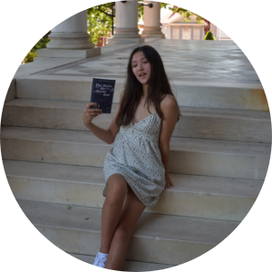 Photo of a young Asian woman in a dress holding a  book sitting on white marble stairs