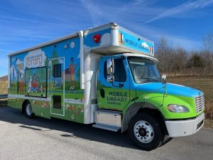 A large truck with Mobile Library on the front and side. The side features an outdoor scene of various people reading, walking, playing music on a sunny day