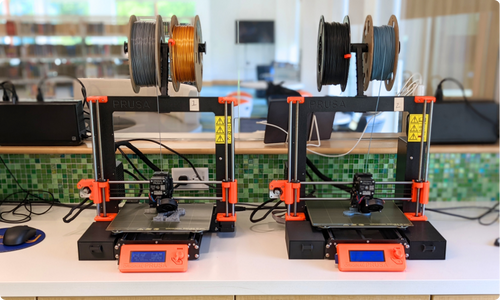 Two orange and black FDM 3D printers sitting next to one another on a counter