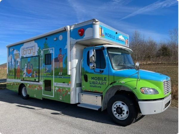 A large truck with Mobile Library on the front and side. The side features an outdoor scene of various people reading, walking, playing music on a sunny day