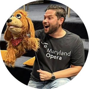 A man in a black Maryland Opera tshirt holds a large dog puppet