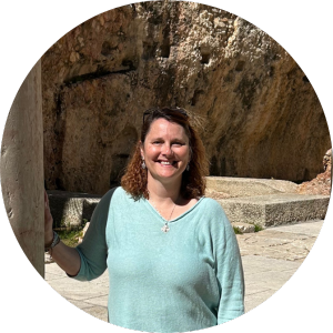 A woman with shoulder length brown hair wearing a light blue shirt stands in front of a rock structure on a sunny day, smiling at the camera