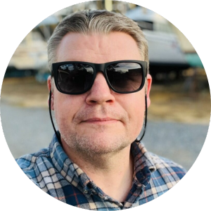 A man with short hair wearing sunglasses and a blue plaid shirt looks at the camera