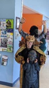A man in a bigfoot mask with his arms raised standing behind a woman in a gold cape and mask and a small child wearing a werewolf mask