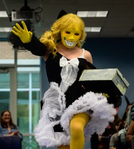 A person in the cosplay contest, dressed as a character with a distinct yellow smiling mask, blonde wig, and a black and white ruffled outfit holding a box.