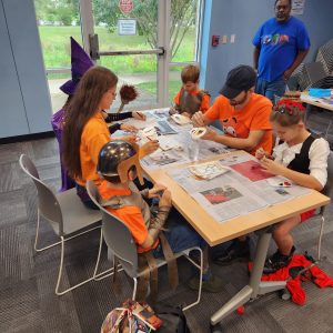 A group of adults and children sit at a table painting masks
