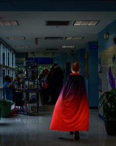 A figure wearing a cloak that fades from red to black walks down a dark hallway with their back to the camera