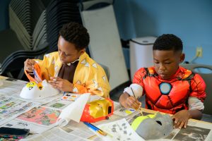 Two young Black children in superhero costumes sit at a table painting masks