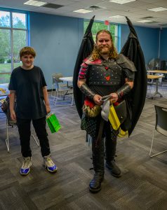 A white man with long hair and a beard wearing a red and black suit of armor and wings smiles at the camera