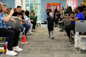 A young child dressed in black with white paint on their face walks down the center aisle in a cosplay contest, past a diverse crowd sitting in chairs on either side