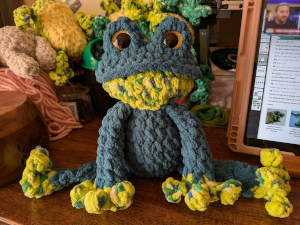 A large crocheted blue and yellow frog with brown eyes sitting on a desk with various colors of yard behind it.