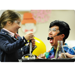 A woman with short brown hair and a young girl with long brown hair, both wearing safety glasses, perform a science experiment