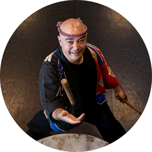 Mark Rooney stands behind a Taiko drum, smiling and tossing a drumstick into the air. He is wearing traditional Japanese dress for drumming.