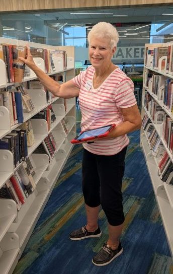 A woman in a red and white striped shirt stands between two shelves of books, holding a tablet and smiling at the camera