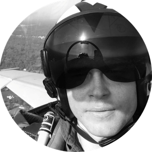 Black and white photo of Zach Fisher wearing a helmet the covers his eyes. The photo was taken inside a plane and the wing is visible in the background.