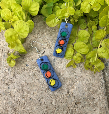 two rectangular gray earrings made of polymer clay decorated with colored circles and black outlines, on a background of stone and green vines