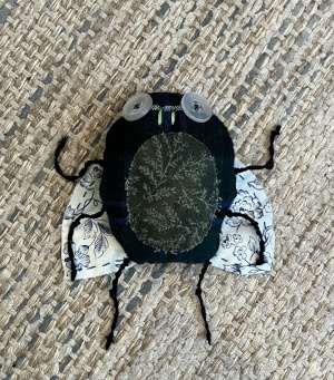 A stuffed fabric fly, constructed out of black and dark green fabric, with white and blue flowered fabric wings, legs made out of knotted black embroidery thread, and eyes of transparent buttons.