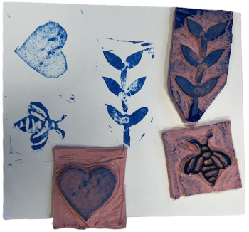 Three hand-carved rubber stamps placed on a square of cardstock with a print from each stamp made with blue ink. The stamps are of a bumblebee, a heart, and a budding plant with six leaves.