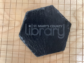 Hexagonal slate coaster with an engraving of the St. Mary's County Library logo
