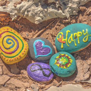 Five small rocks painted with various designs and colors, including a purple heart, an infinity symbol, and the word Happy