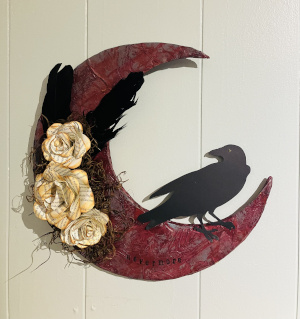 A red textured crescent moon with the silhouette of a crow sitting within the crescent, to the right. Two black feathers, dried grass, and sepia colored book page flowers adorn the left outer side of the moon. The moon sits on a white planked background.