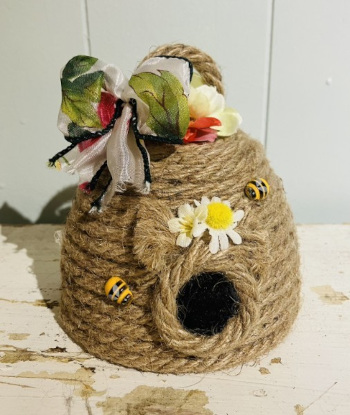 A beehive crafted out of rope with artificial flowers and ribbons topping it and artificial bees and daisies surrounding the opening.