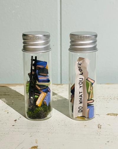 Two small glass bottles with tiny books inside. One also has a ladder, and the other a banner reading Do What You Love
