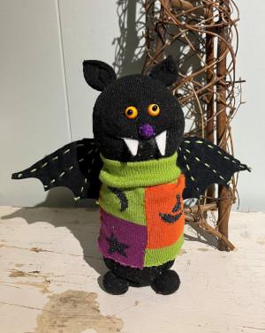A stuffed animal bat with yellow eyes and white fangs, wearing a green, purple, and orange sweater