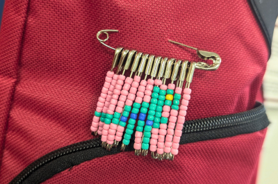 Small, brightly colored beads are strung onto safety pins, creating the pattern of a dinosaur. The safety pins hang vertically from one large safety pin, creating a dinosaur beaded brooch.