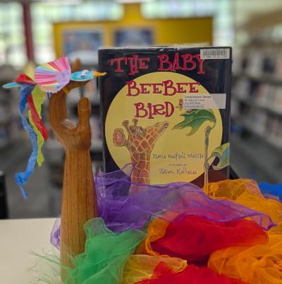 Picture book The Baby BeeBee Bird standing behind a pile of colorful fabric. A wooden arm stands next to the book holding a small bird crafted out of paper
