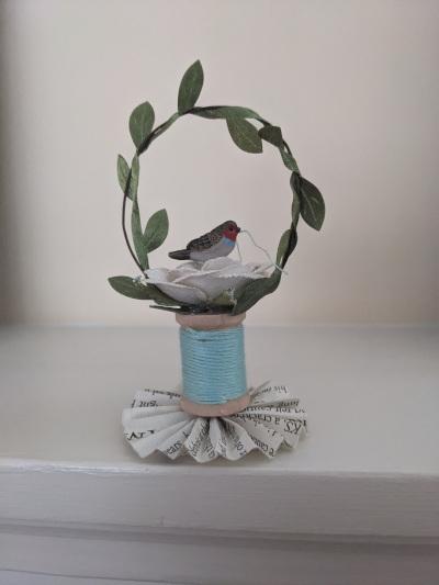 A wooden spool with blue thread holding a bird in a bird's nest with a wreath of green leaves