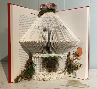 An open book with pages folded down in a pattern to create the shape of a birdcage. It is embellished at the top and bottom with paper flowers, lace, and moss with a small orange paper bird to the bottom right.