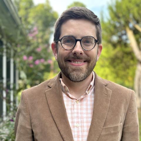 A man with dark hair and beard and mustache wearing glasses and a brown jacket stands in front of trees, smiling at the camera