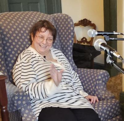 A woman with short brown hair wearing glasses and a black and white shirt sits smiling in a blue chair surrounded by three microphones
