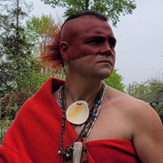 An Indigenous man with red face paint and draped in red fabric looks to the right of the camera