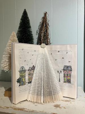 An open book standing on its bottom edge, with the middle pages folded to resemble a tree shape. Three decorative trees are in the background.