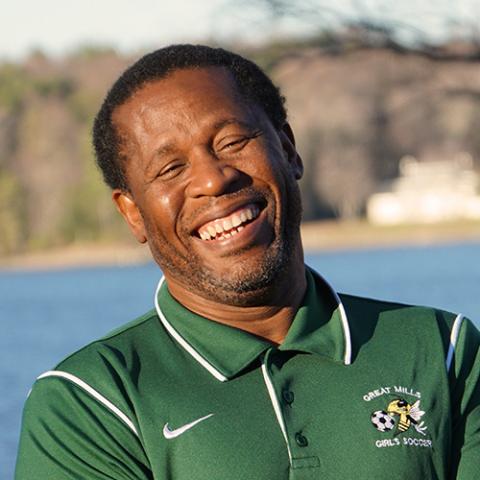 A Black man in a green polo shirt stands laughing in front of a body of water
