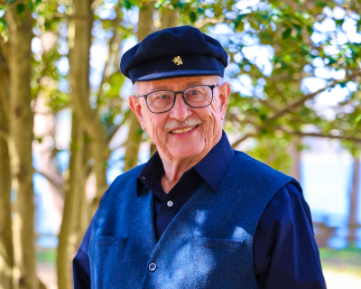 A man wearing glasses, a blue sweater vest, and a blue cap smiles at the camera