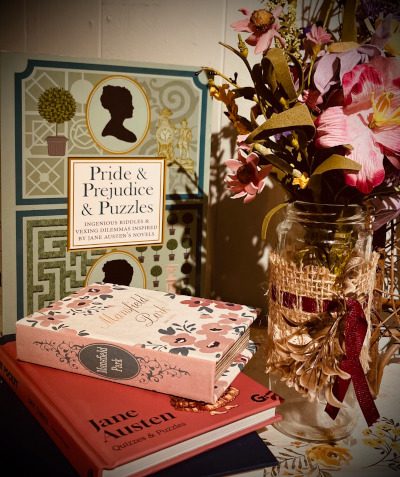 Two Jane Austen books stacked on a table next to a vase full of pink flowers. A Pride & Prejudice puzzle box stands in the background.