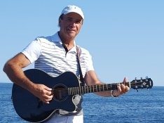 A man in a white striped polo shirt and hat stands in front of a body of water holding a guitar